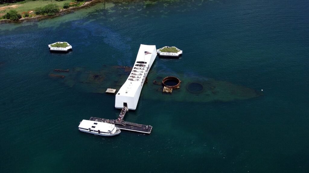 An aerial view of the USS Arizona Memorial at Pearl Harbor, with the outline of the sunken battleship visible beneath the water.